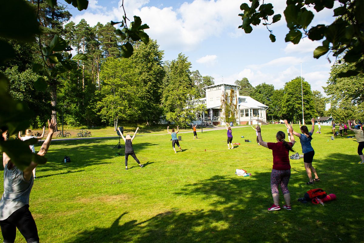 A dozen people exercising on the grass in a park, a villa in the background.