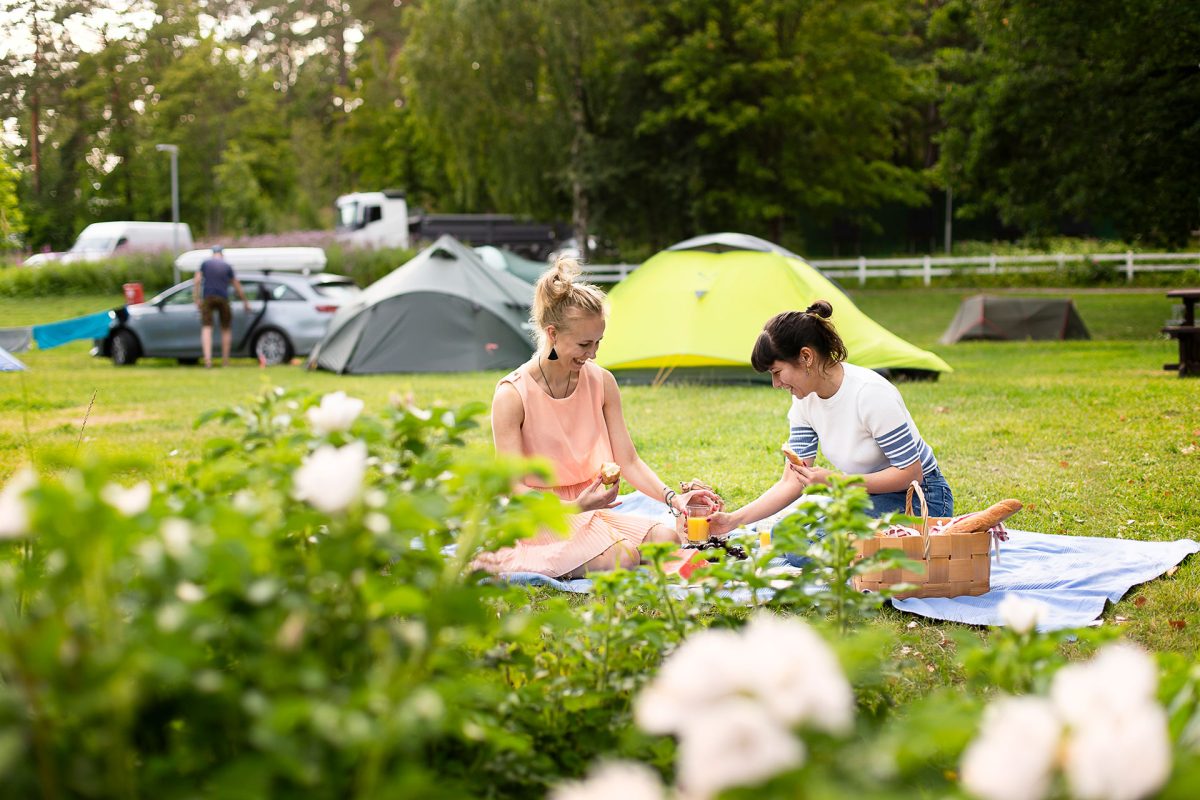 Telttoja näkyy leirintäalueen nurmikolla, kaksi henkilöä ovat piknikillä.