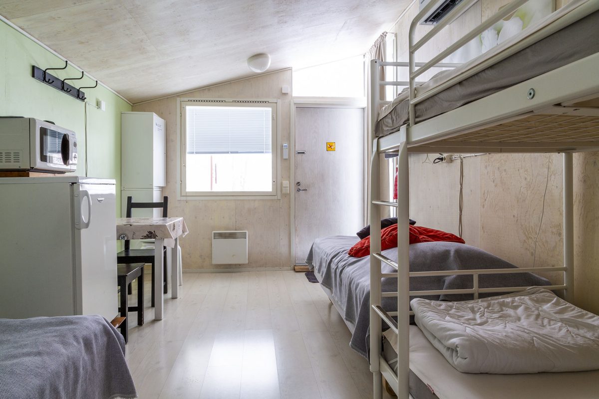 Stuginteriör, en våningssäng och två vanliga sängar, i bakgrunden kylskåp, mikrovågsugn och matbord för två personer.