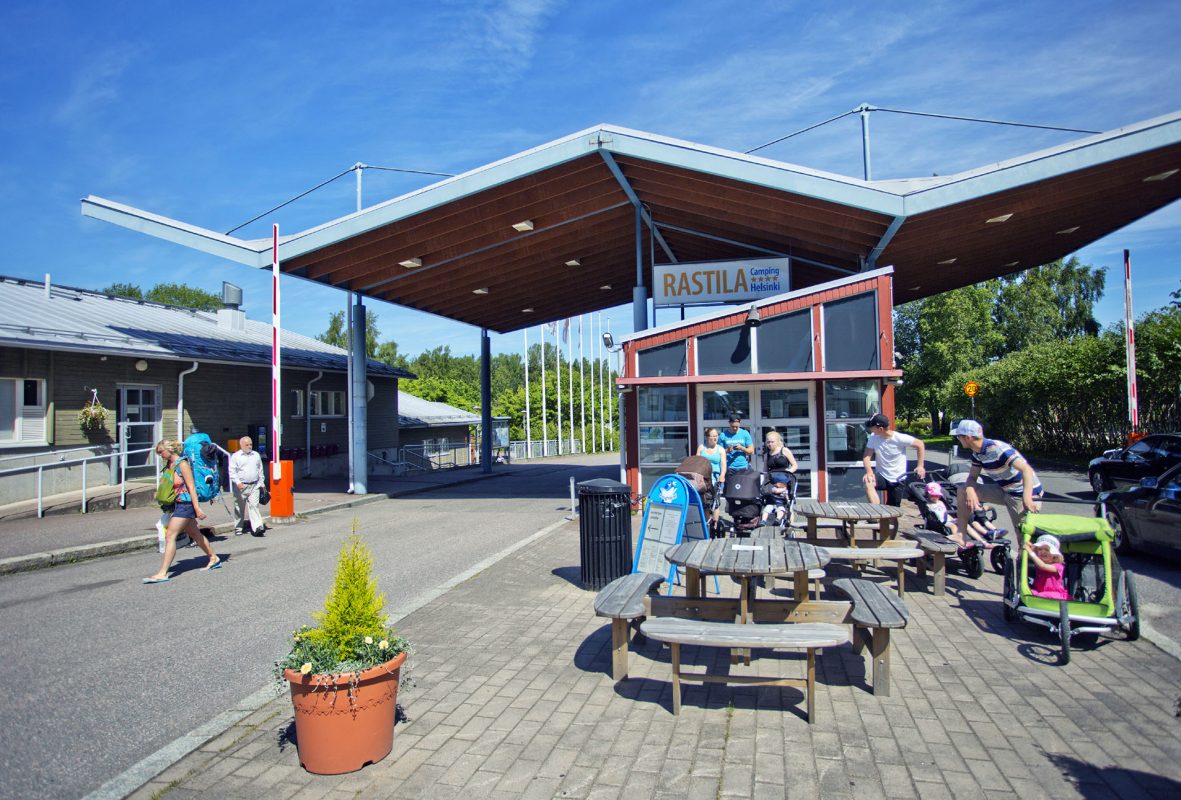 Front gate and reception of the camping site, tables, chairs and a group of travellers with a pram in the background.