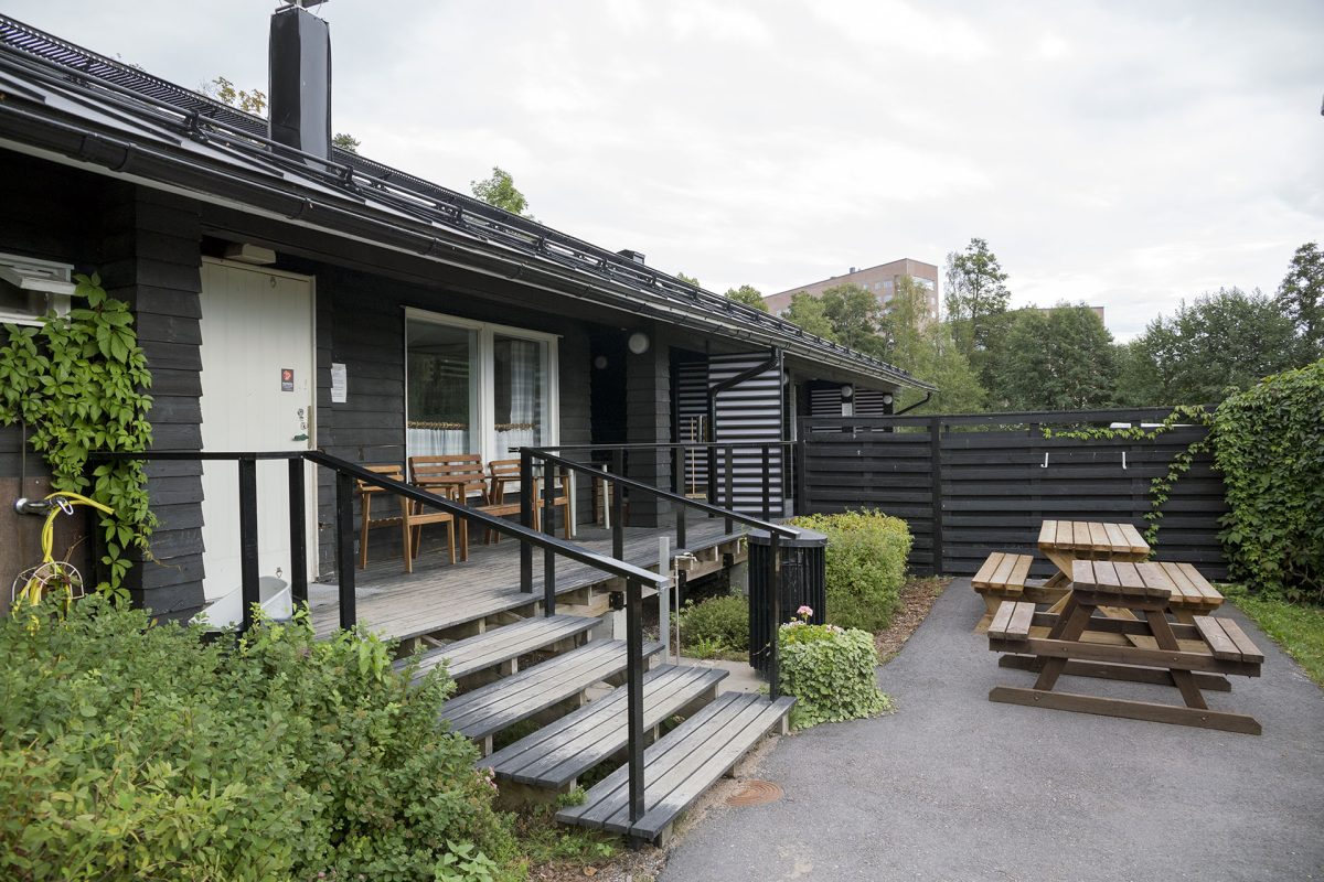 Sauna building from the outside, sauna terrace with chairs, tables and benches in front of the building, a wooden fence separating the saunas in the background.